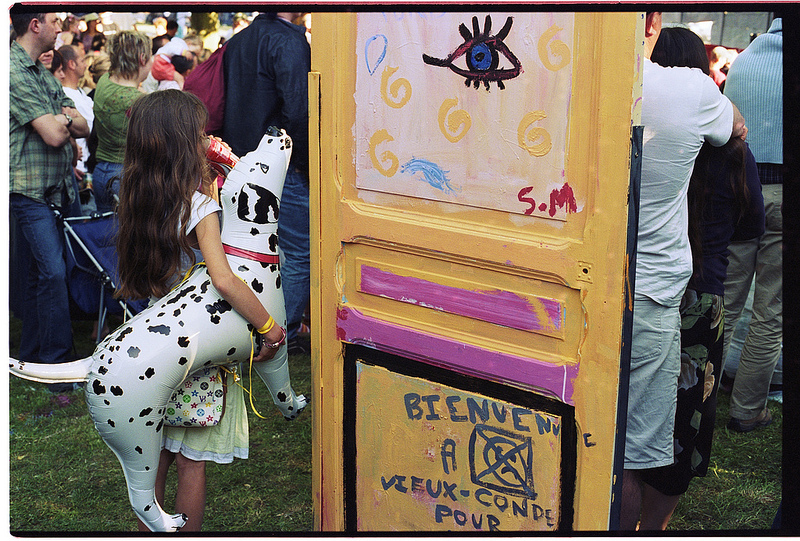 Une fille tenant un chien en plastique se tient devant une porte colorée avec des graffitis, dans une foule en arrière-plan.