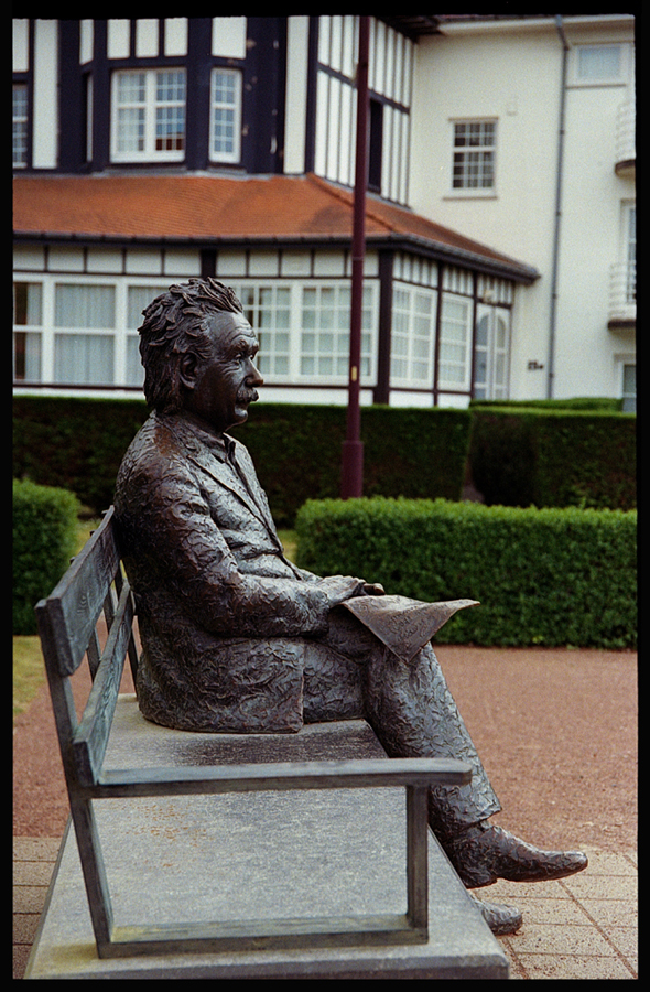 Statue en bronze d'un homme assis sur un banc, semblant lire, avec un bâtiment en arrière-plan.