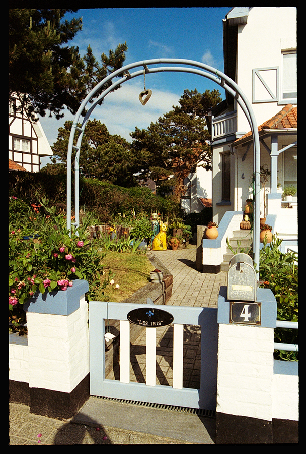 Entrée d'une maison avec une arche décorative, un jardin fleuri et des poteries artistiques visibles en arrière-plan.