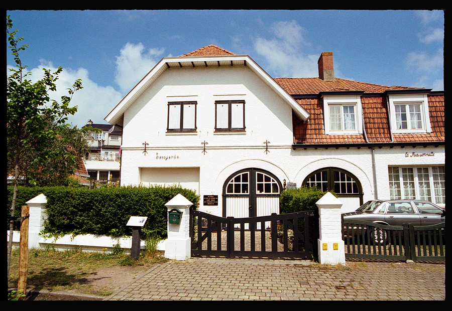 Vue d'une maison élégante à De Haan, Belgique, avec un style architectural typique, un jardin et une allée pavée, sous un ciel bleu partiellement nuageux.