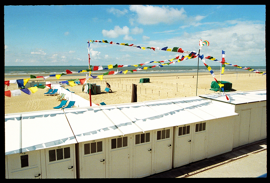 Vue d'une plage à De Haan avec des cabines de bain blanches et des chaises longues colorées. Des fanions multicolores sont suspendus au-dessus du sable, avec la mer en arrière-plan et un ciel partiellement nuageux.