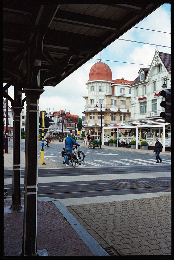 Vue d'une rue animée à De Haan, montrant des bâtiments d'architecture élégante, des personnes se déplaçant, et des panneaux de signalisation.