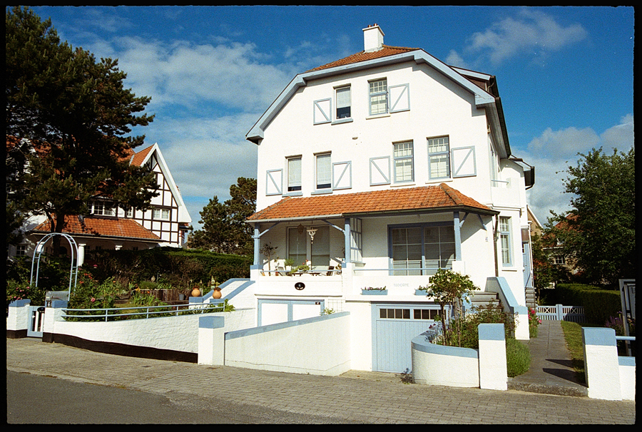 Maison typique de De Haan avec façade blanche et toit en tuiles, entourée de verdure et d'une allée pavée sous un ciel ensoleillé.