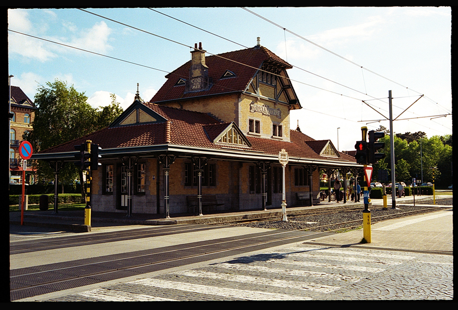 Vue de l'ancienne gare de De Haan, Belgique, avec son architecture typique en briques, toiture en tuiles et éléments décoratifs, entourée de verdure et de rues pavées.