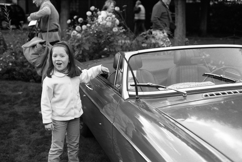 Une jeune fille souriante se tient à côté d'une voiture décapotable, avec des fleurs en arrière-plan et des passants. La photo est en noir et blanc.