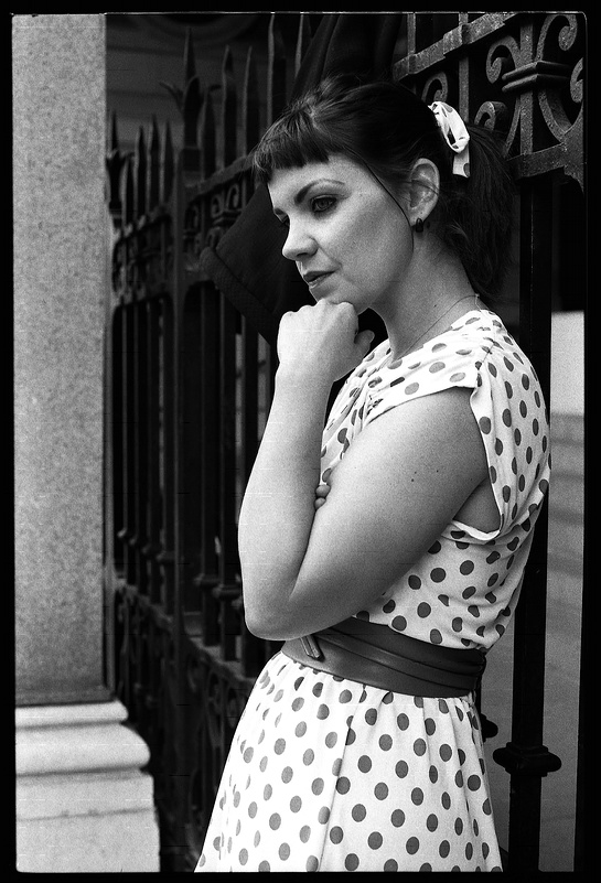 Portrait d'une femme pensive avec une robe à pois, en noir et blanc, se tenant près d'une clôture en fer forgé.