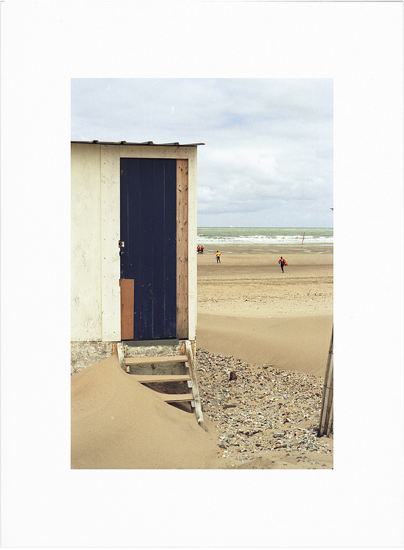 Un cabanon de plage avec une porte bleue, des marches en bois et une plage de sable en arrière-plan, quelques personnes au loin et un ciel nuageux.