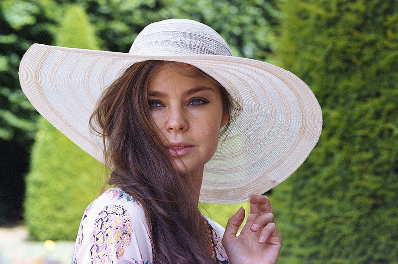 Portrait d'une femme portant un grand chapeau en paille, avec des cheveux bruns et des yeux clairs, dans un environnement verdoyant.