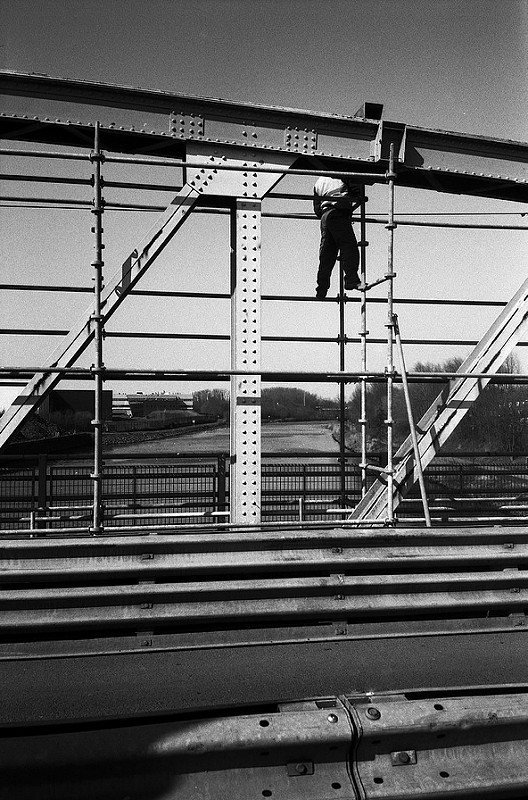Un homme travaille en hauteur sur une structure en métal d'un pont, utilisant un échafaudage, avec un paysage de canal en arrière-plan.