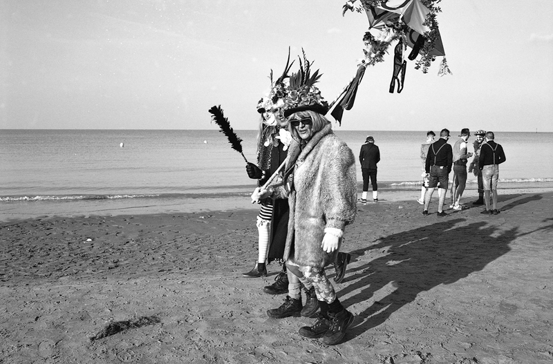 Deux personnes déguisées marchant sur la plage, l'une portant un long manteau et des accessoires colorés, tandis que d'autres sont visibles en arrière-plan.
