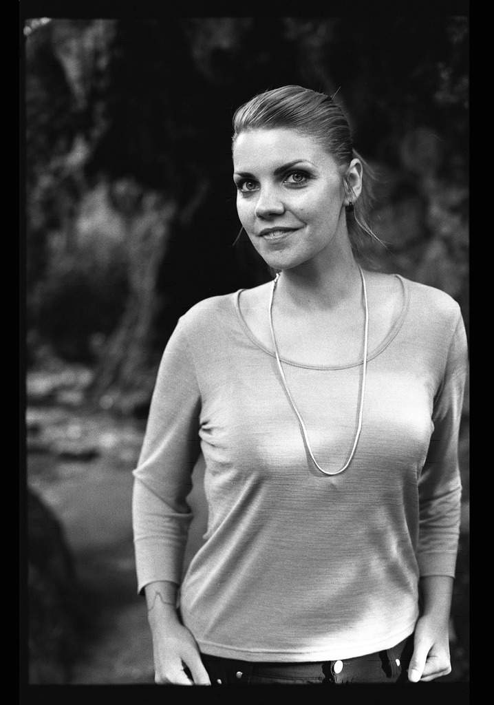 Portrait en noir et blanc d'une femme avec un haut clair. Elle a les cheveux longs et attachés, et elle se tient debout avec un léger sourire.