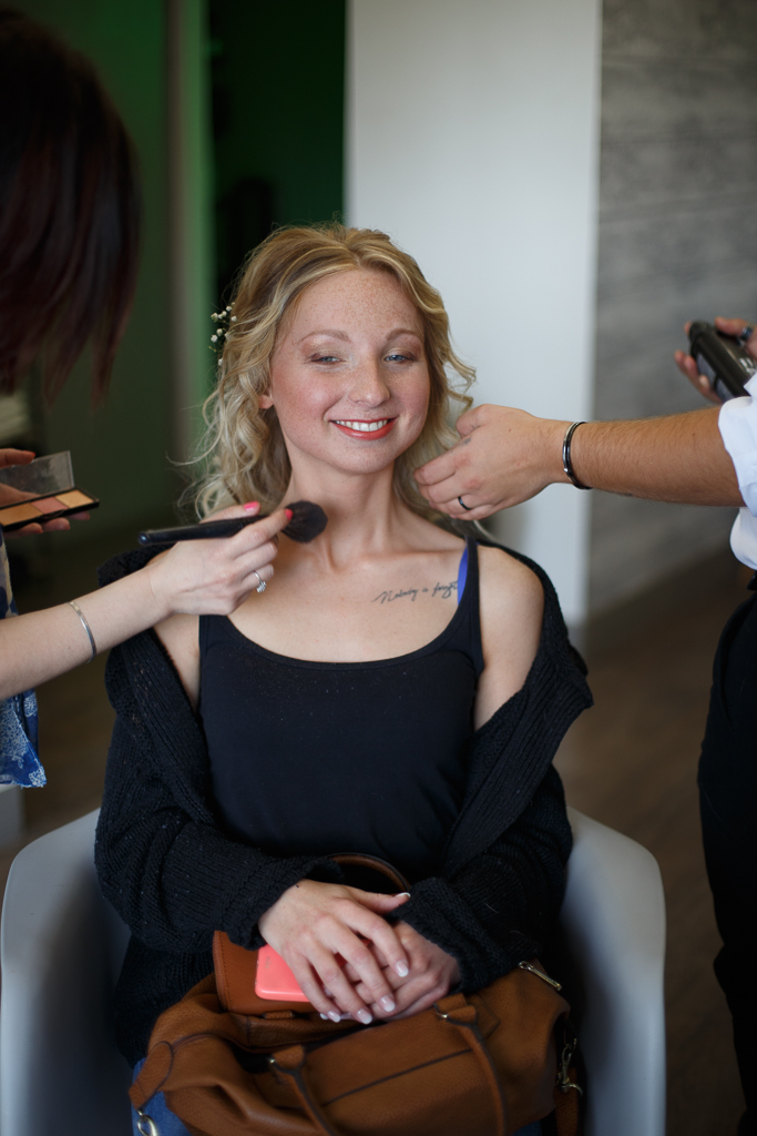 Une mariée assise dans un fauteuil se fait maquiller par deux personnes, avec un fond moderne et lumineux.