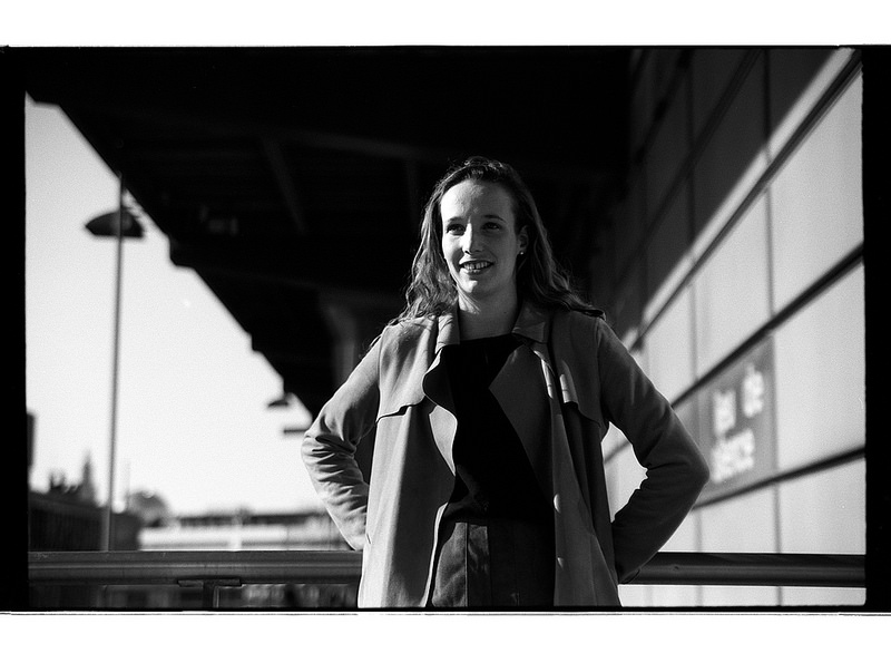 Portrait en noir et blanc d'une femme souriante debout, vêtue d'un manteau, avec un éclairage contrasté mettant en valeur les ombres et la lumière.