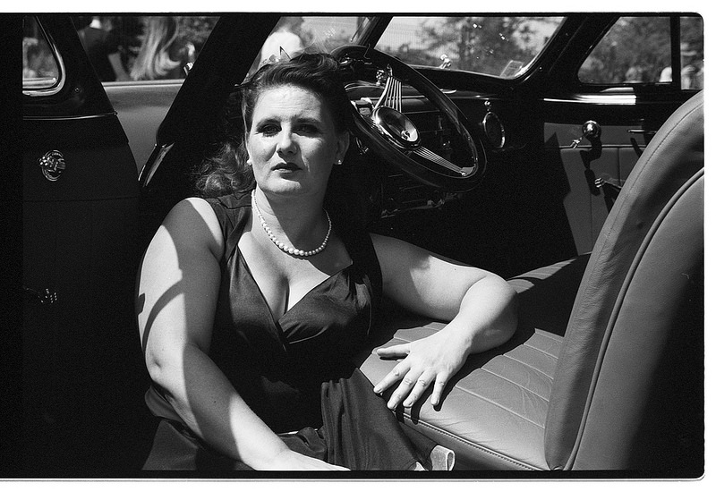 Une femme assise à l'intérieur d'une voiture classique, vêtue d'une robe noire et de perles, avec un éclairage contrasté en noir et blanc.