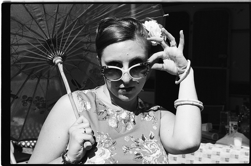 Une femme posant avec un parasol et des lunettes de soleil, habillée d'une robe à fleurs, en noir et blanc.