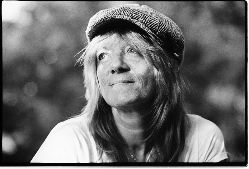 Portrait en noir et blanc d'une femme portant une casquette, regardant vers le haut avec un léger sourire, dans un environnement lumineux flou en arrière-plan.