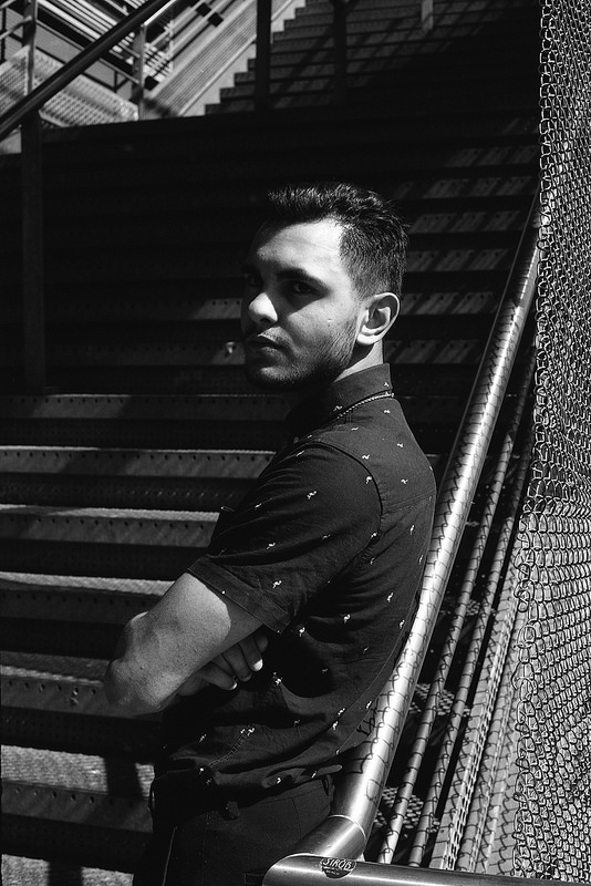 Un homme se tenant sur des escaliers, vêtu d'une chemise noire à motifs, dans un éclairage fort qui crée des contrastes marqués en noir et blanc.