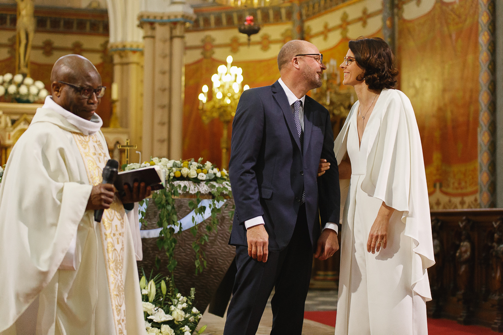 Un couple souriant échange des regards pendant une cérémonie religieuse, avec un prêtre tenant un livre de prières en arrière-plan.