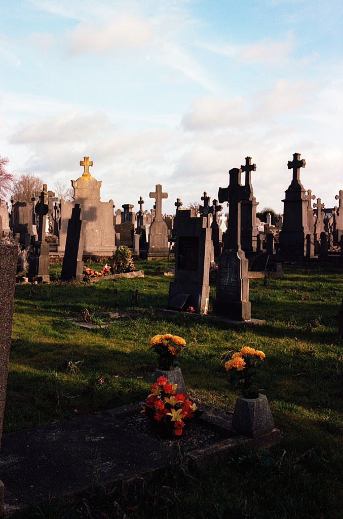 Vue d'un cimetière avec des pierres tombales et des fleurs colorées sur certaines tombes, sous un ciel partiellement nuageux.