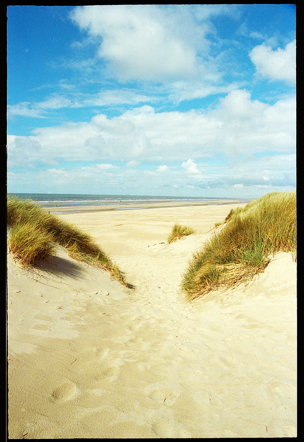 Vue sur une plage de De Haan avec des dunes de sable et un ciel partiellement nuageux.