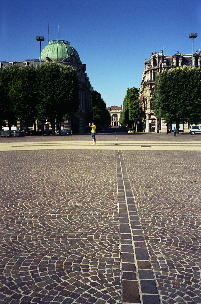Un homme portant un t-shirt jaune se tient au centre d'une place bordée d'arbres, avec des bâtiments historiques en arrière-plan et un ciel bleu dégagé.