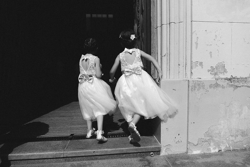 Deux jeunes filles en robes blanches courent ensemble vers une porte, capturant un moment de mouvement et de joie en noir et blanc.