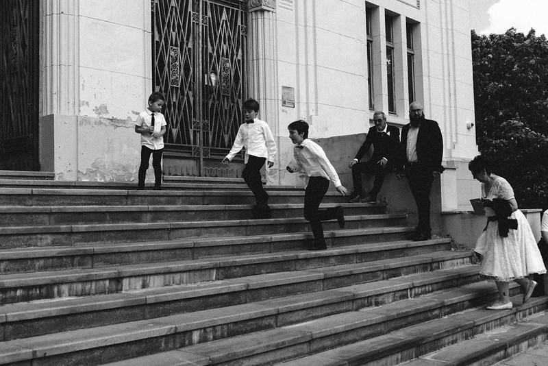 Trois enfants en costume descendant les escaliers, avec des adultes assis au fond, dans une scène animée en noir et blanc.