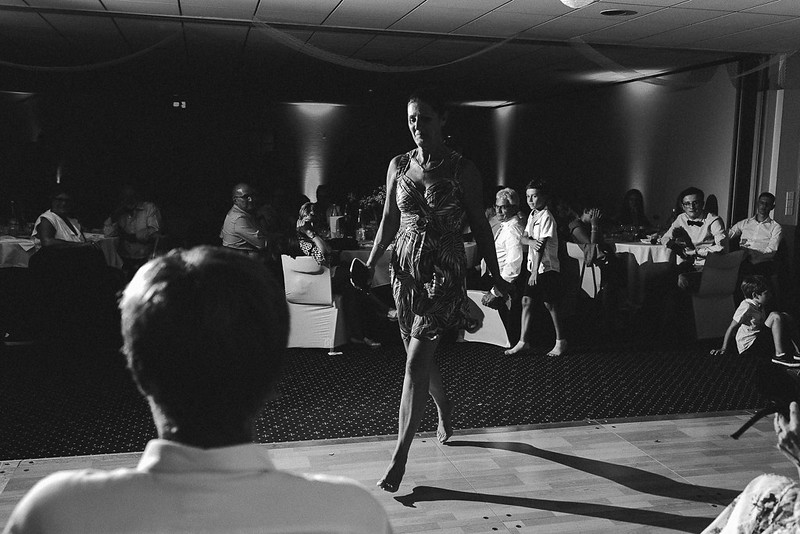 Une femme marche sur la piste de danse lors d'un événement, tandis que des invités l'observent à table. La photo est en noir et blanc, capturant l'énergie du moment et les interactions des participants.