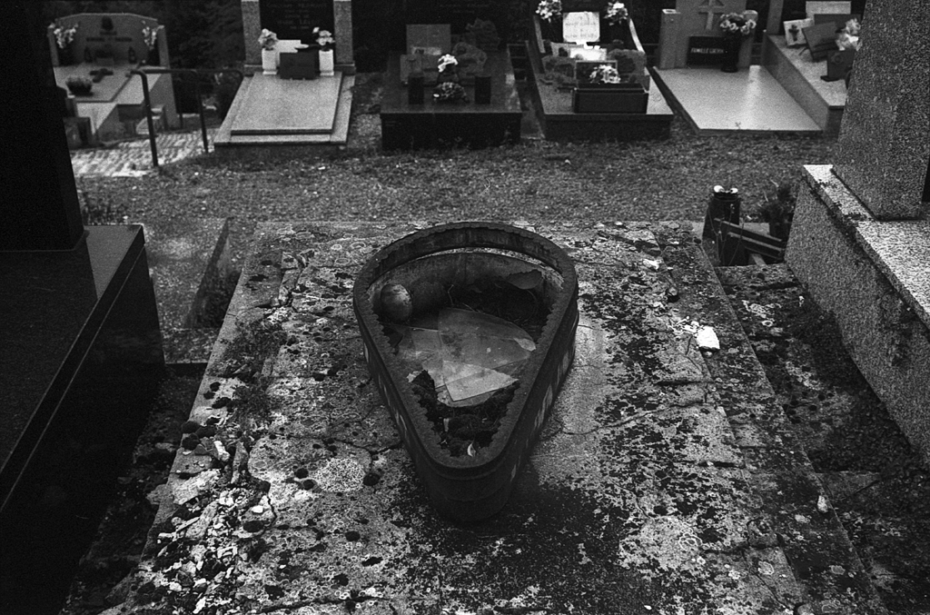 Vue d'une tombe dans un cimetière, avec un vase cassé sur une pierre tombale recouverte de lichen et de débris, en noir et blanc.