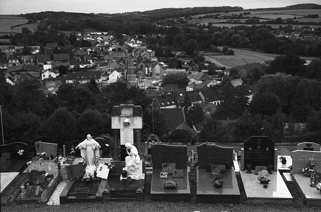 Vue d'un cimetière avec des tombes et des statues, surplombant une ville et des champs au loin, photographiée en noir et blanc.
