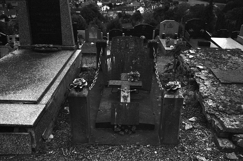 Cimetière avec une tombe ancienne, entourée de gravillons et de fleurs, sous un ciel nuageux.
