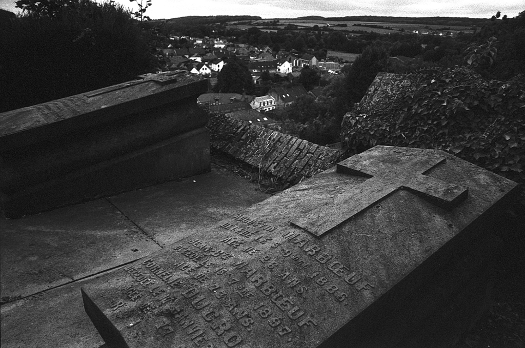 Vue d'un cimetière avec une pierre tombale gravée et une croix, en arrière-plan un paysage de collines et de maisons. Photographie en noir et blanc.
