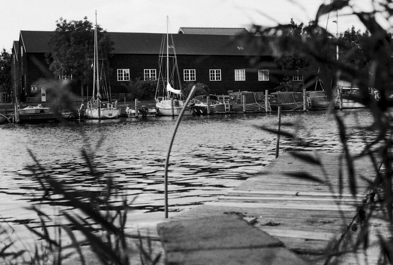 Vue en noir et blanc d'un port avec des bateaux amarrés, un quai en bois au premier plan et un bâtiment en arrière-plan, le tout entouré de végétation.
