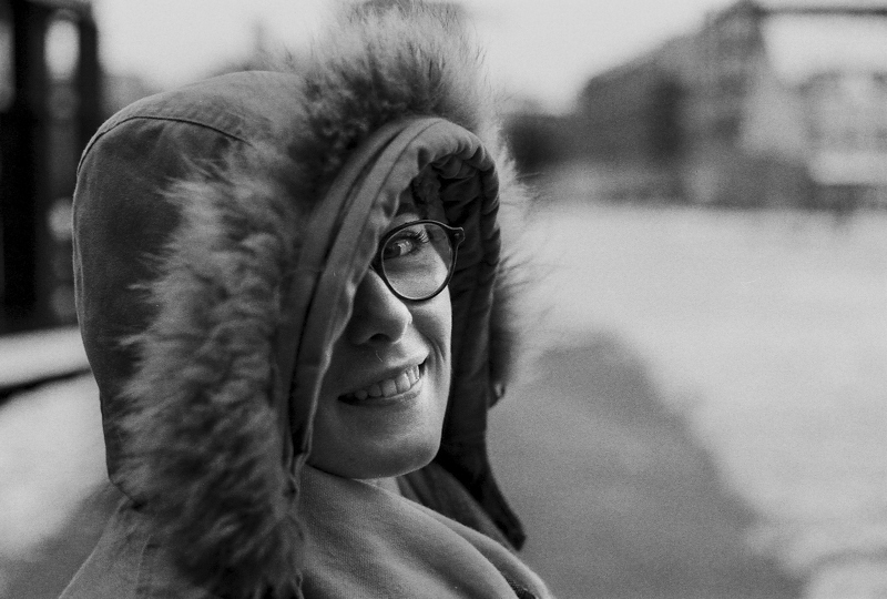 Portrait d'une femme souriante portant un manteau à capuche, en noir et blanc, sur un fond d'ambiance urbaine.