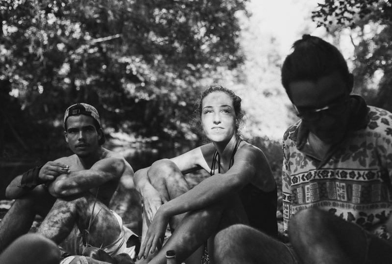 Groupe de trois personnes assises près d'une rivière, en noir et blanc, avec des textures de lumière et d'ombre accentuées.