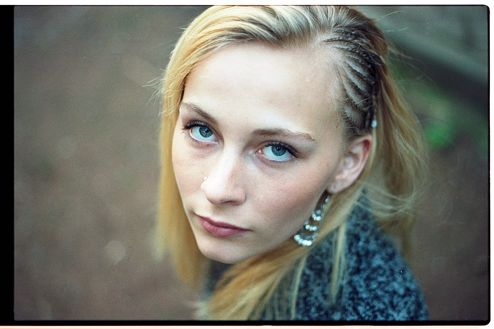 Portrait d'une jeune femme avec des cheveux blonds, portant des boucles d'oreilles, fixant l'objectif avec un regard intense, pris dans un environnement naturel flou.