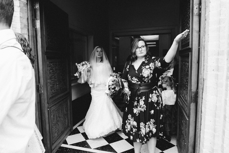 Une mariée en robe blanche sort d'une porte avec un bouquet à la main, tandis qu'une femme en robe noire à fleurs lui fait signe de la main, dans un cadre intérieur au sol en damier.