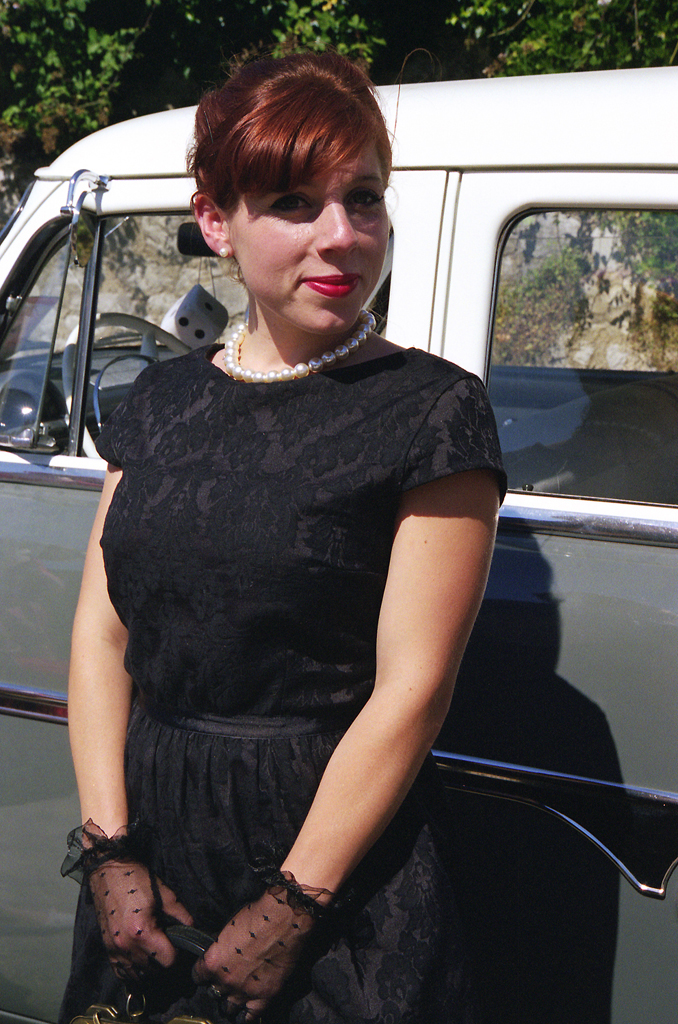 Portrait d'une femme avec des cheveux roux, portant une robe noire et des gants en dentelle, devant une voiture classique.