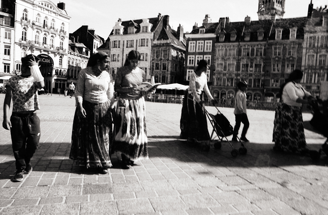 Une scène de rue en noir et blanc montrant des personnes marchant sur une place à Lille, avec des bâtiments historiques en arrière-plan.