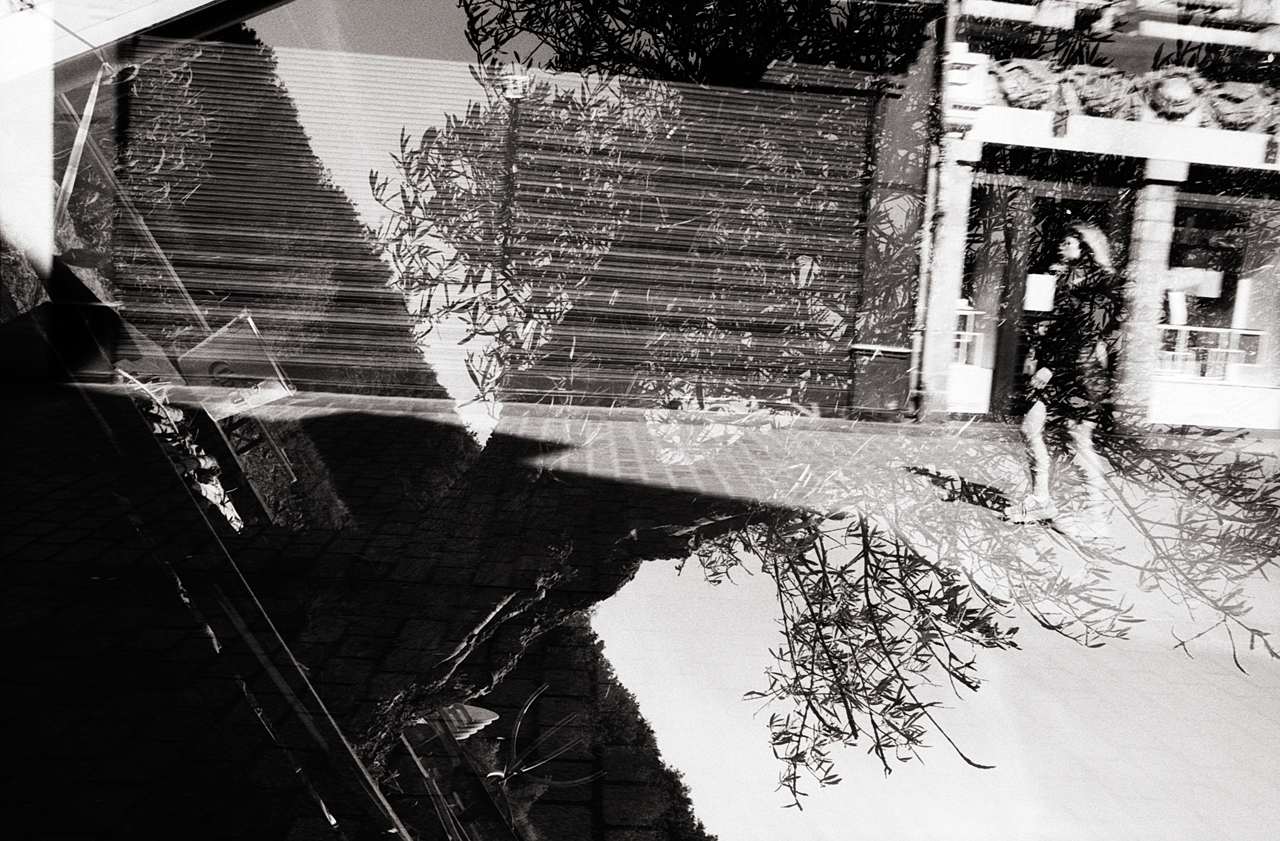 Double exposition en noir et blanc montrant des reflets d'arbres et une silhouette humaine dans une rue.