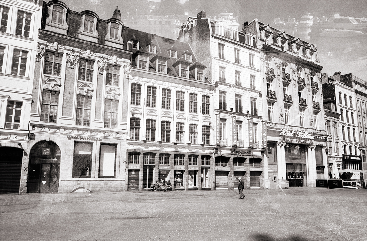 Photographie en noir et blanc représentant une place à Lille, avec des bâtiments historiques et une personne marchant au centre.