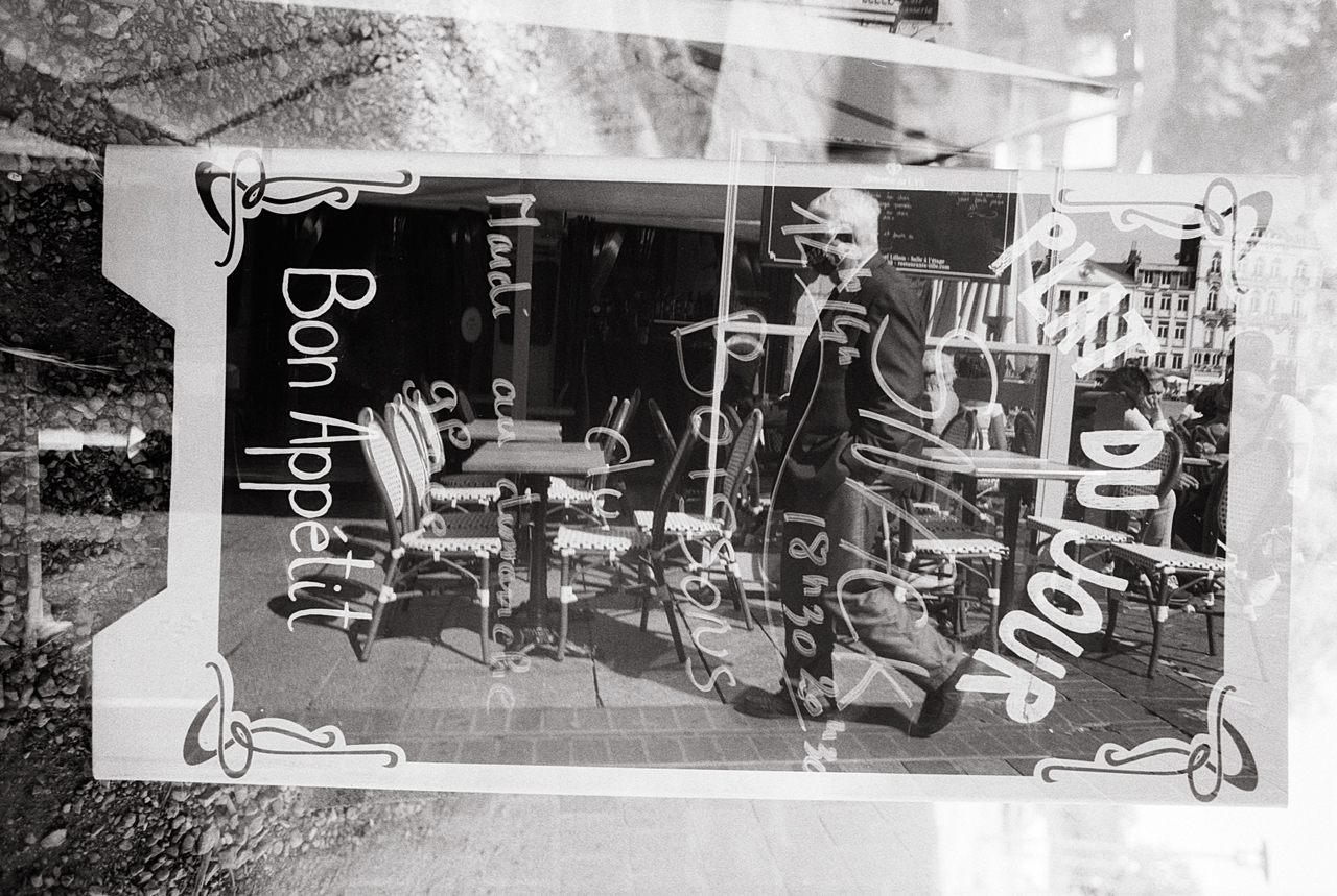 Double exposition argentique d'une scène de rue avec un panneau 'Bon Appétit' en noir et blanc, incluant des silhouettes floues et des détails de chaises de café.
