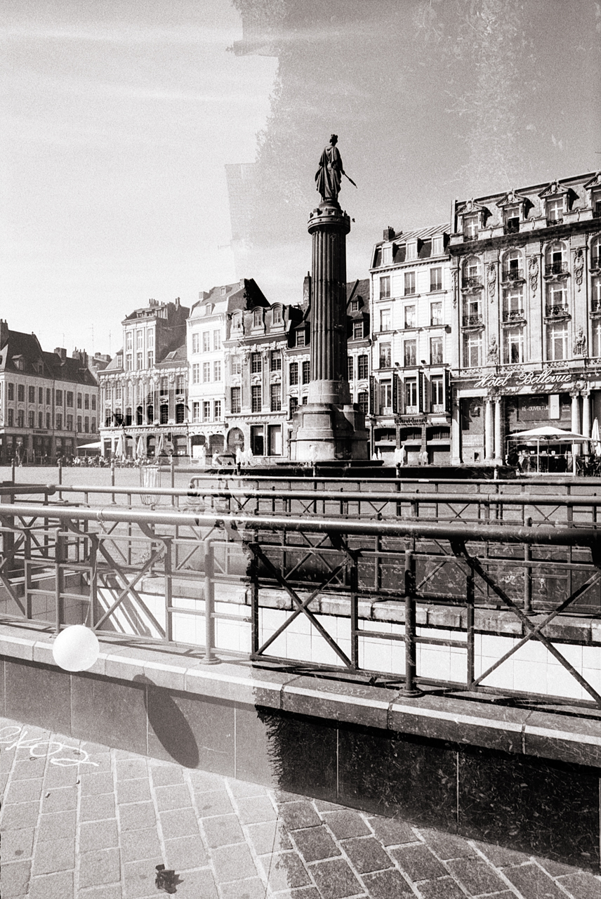 Double exposition en noir et blanc d'une place à Lille, montrant un monument et des bâtiments historiques avec un effet de superposition.