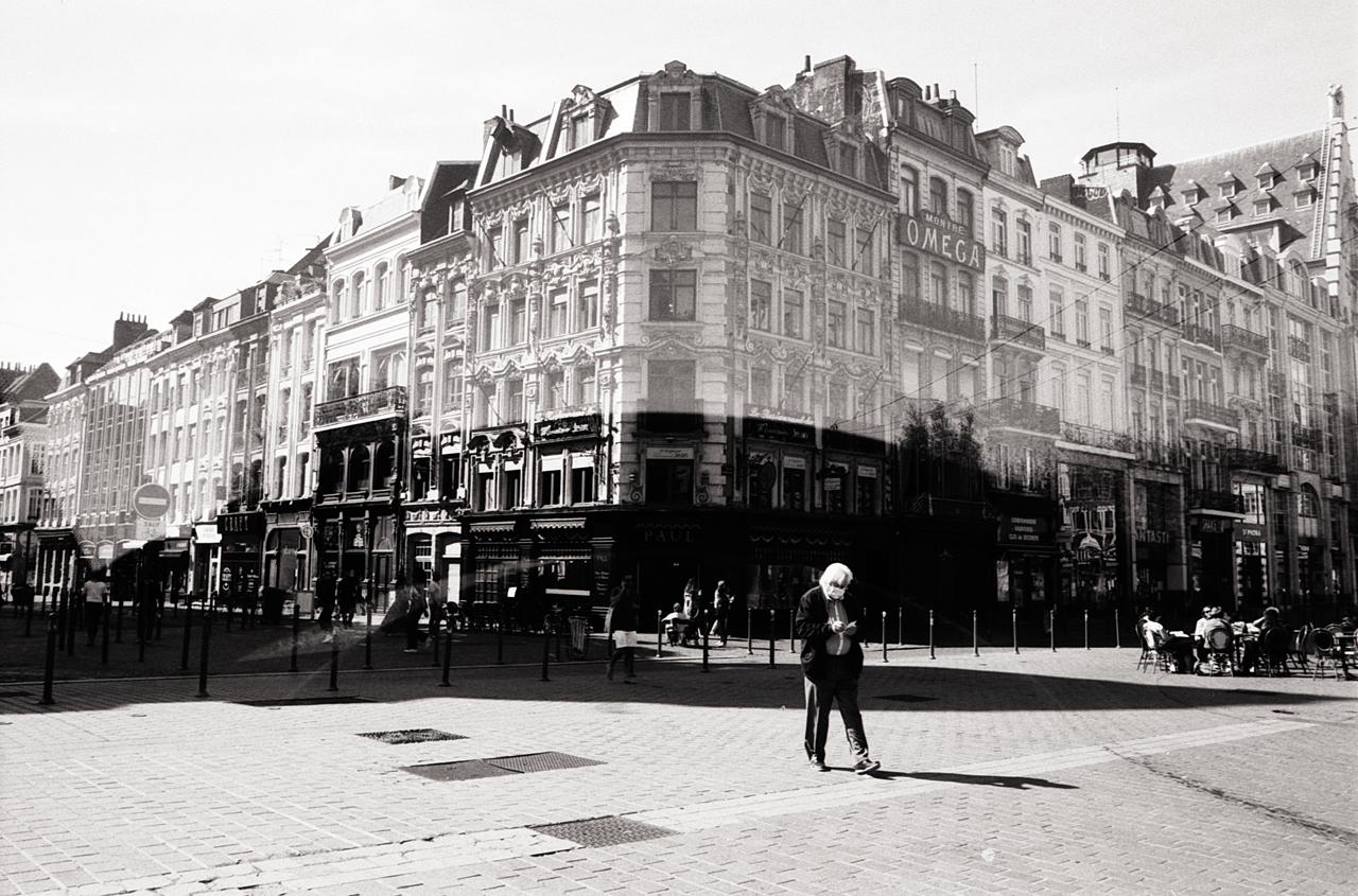 Photographie en noir et blanc d'une rue animée à Lille, avec des bâtiments historiques en arrière-plan et des passants flous en raison d'une double exposition.