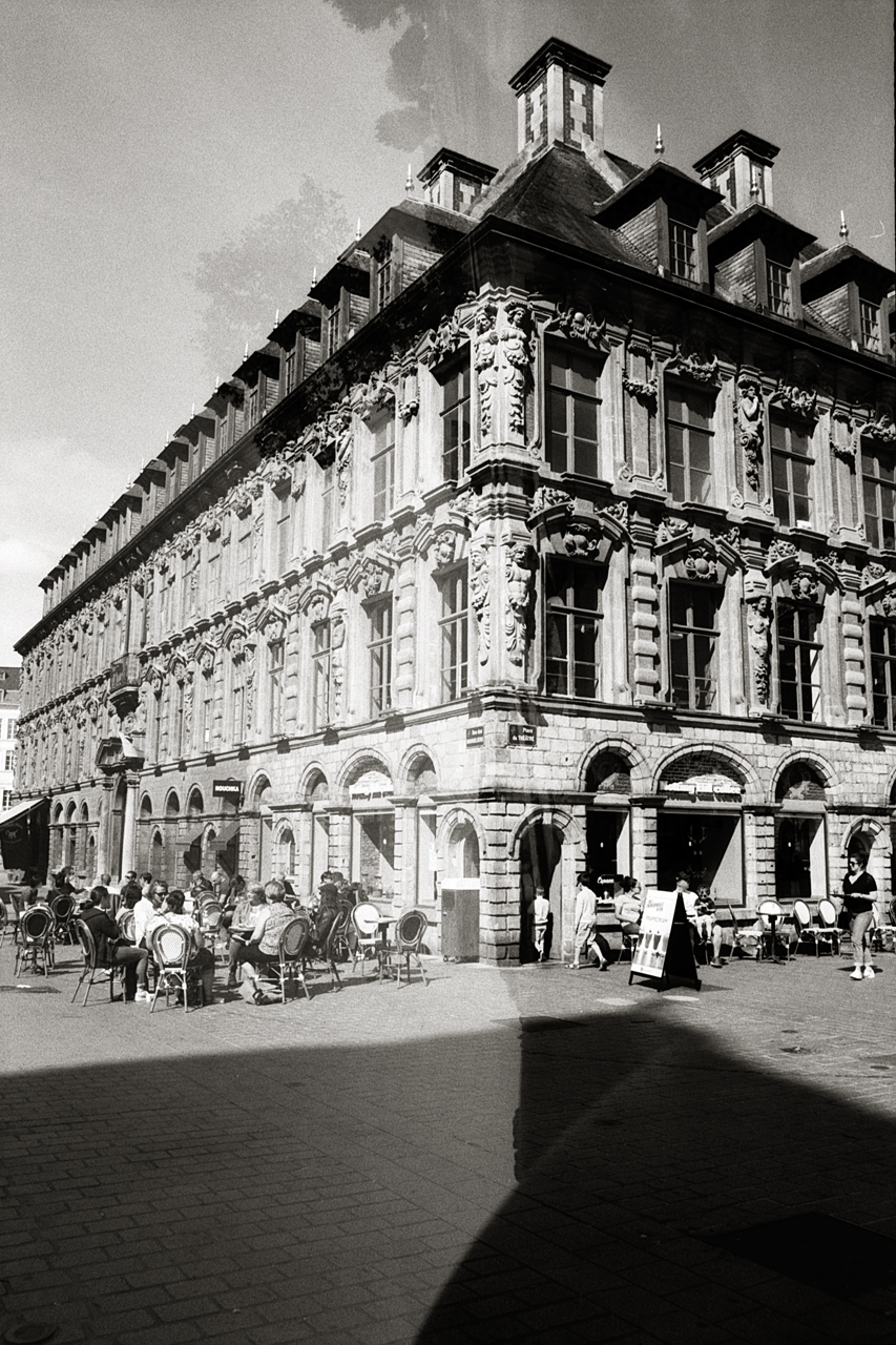 Photographie en noir et blanc d'un bâtiment à Lille, avec une double exposition montrant une superposition d'éléments architecturaux et des personnes assises en terrasse.
