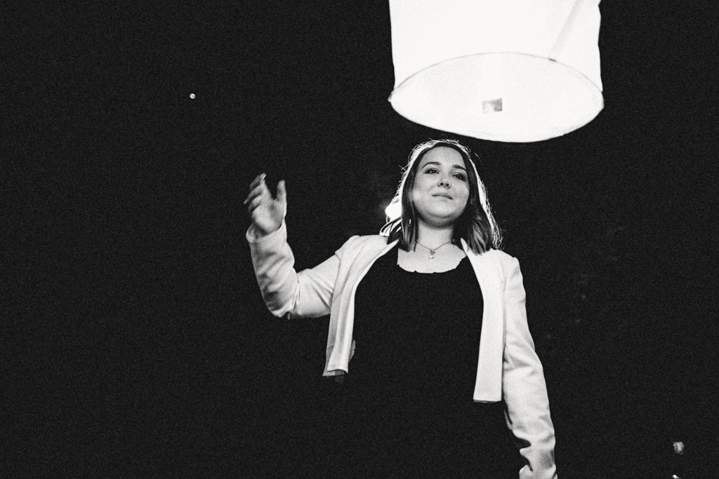 Une femme souriante vêtue d'une veste blanche se tient sous une lanterne flottante, capturée en noir et blanc dans une ambiance nocturne.