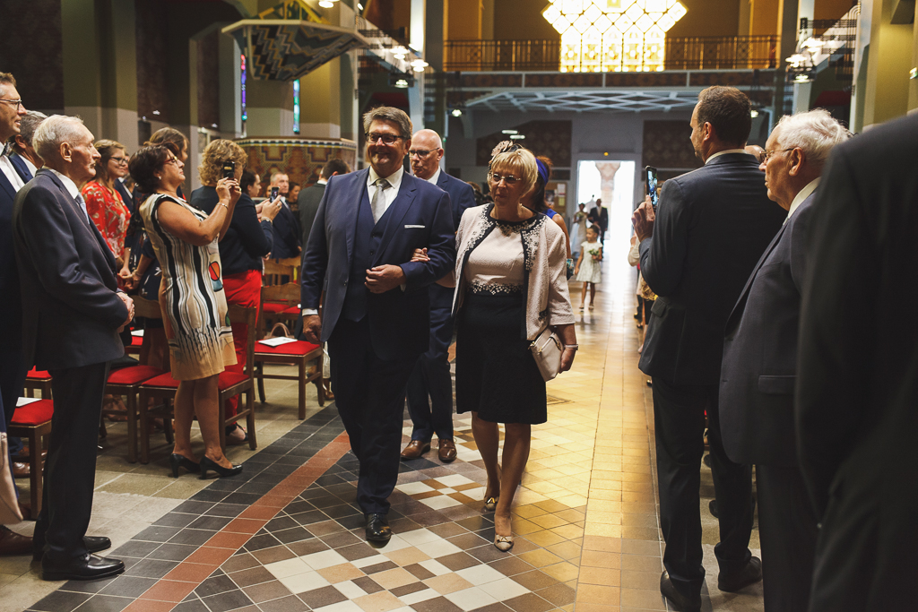 Un couple marchant ensemble dans une église lors d'une cérémonie, entouré de personnes en costume et en tenue élégante, avec des bancs et un éclairage chaleureux.
