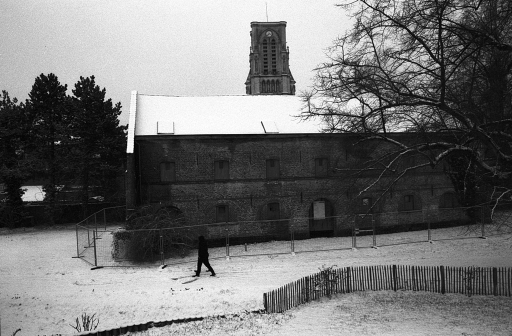 Un homme marchant sur un chemin enneigé, devant un bâtiment en pierre avec un toit recouvert de neige et un clocher visible en arrière-plan, sous un ciel nuageux.