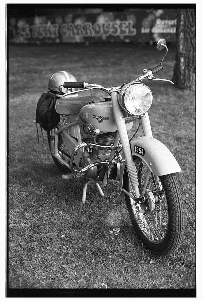 Une moto vintage garée sur l'herbe, avec un casque posé sur le guidon et des couleurs en noir et blanc.