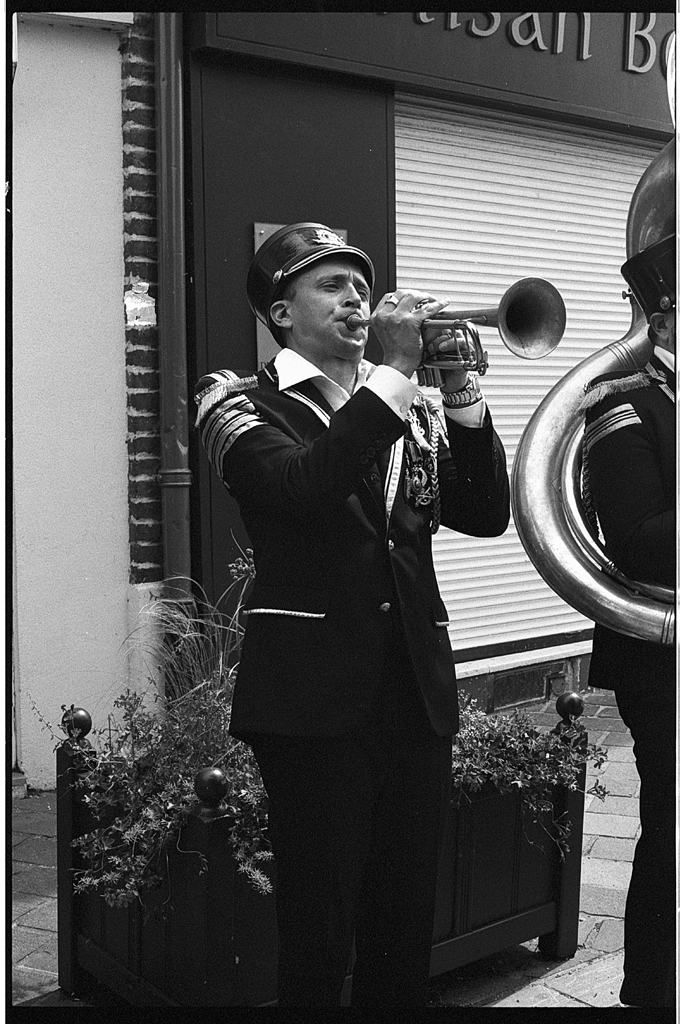 Un musicien en uniforme joue de la trompette lors d'un événement en extérieur, entouré de fleurs dans des jardinières.
